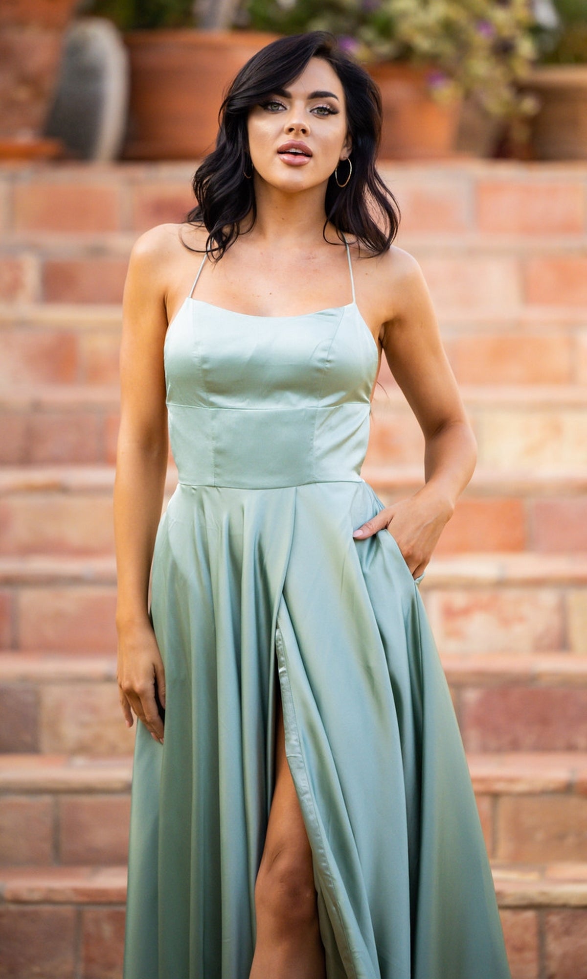 A woman in a Velvi Long Satin Prom Dress Liliana, light green and sleeveless, stands outdoors by stone steps with one hand in her pocket and the other at her side.